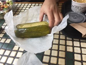 A hand reaches on a black and white tiled table to pick up a pickle. 