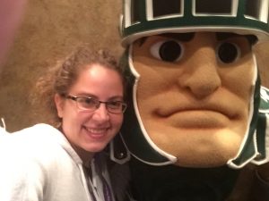 A woman wearing glasses and a white hoodie smiles next to fierce looking Spartan mascot in an elevator.